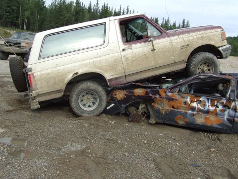 1990 Ford Bronco - A 1990 Ford Bronco is parked on top of a wrecked car in a dirt lot.