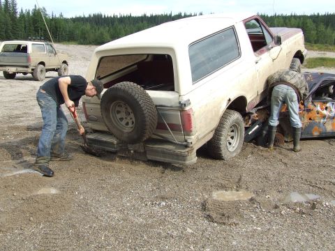 1990 Ford Bronco - Two people attempt to free a muddy 1990 Ford Bronco, with another vehicle visible in the background.