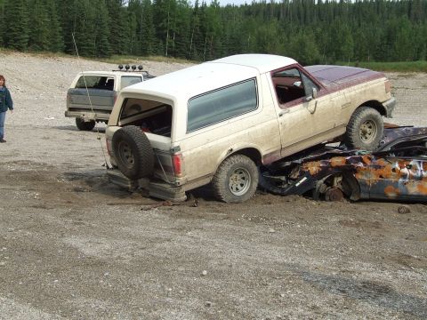 1990 Ford Bronco - A tan and maroon Ford Bronco is precariously perched on a pile of wrecked vehicles in a junkyard.