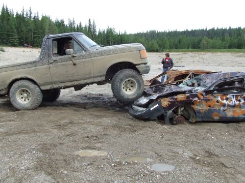 1990 Ford Bronco - 1990 Ford Bronco crushing a wrecked car in a muddy outdoor scene.