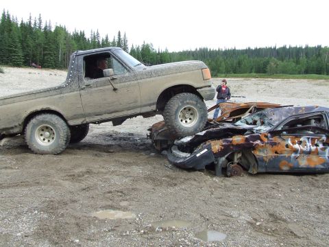 1990 Ford Bronco - A lifted pickup truck is positioned over two heavily damaged and burnt-out cars in a dirt lot.