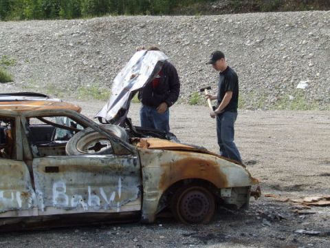 1988 Ford Bronco - Two people examine a heavily damaged car with its hood propped open.