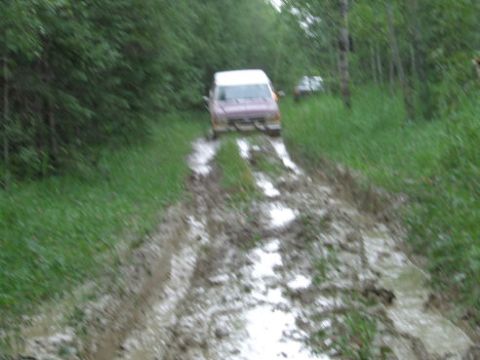 1988 Ford Bronco - A pink van navigates a challenging muddy track through a wooded area, with another vehicle visible ahead.