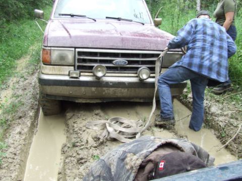1988 Ford Bronco - A dark red Ford truck sits axle-deep in a muddy rut while a person in a blue plaid shirt and jeans hooks a tow strap to the front bumper; a…