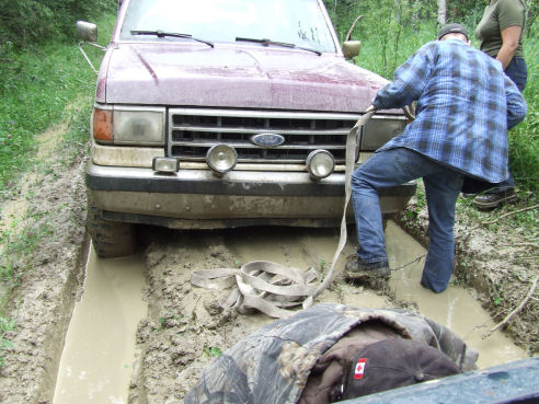 Person rigging a tow strap to a dark red Ford truck stuck axle-deep in mud