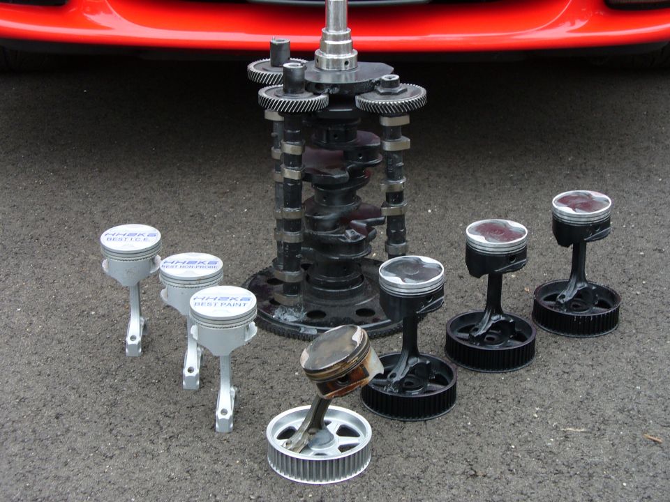 Engine crankshaft, camshafts, and pistons displayed on pavement with a red car bumper in the background. Some pistons have 'HH2KB BEST PAINT' text