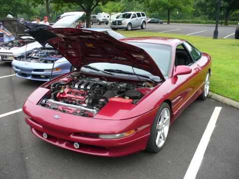 1991 Ford Probe - A red 1991 Ford Probe is displayed with its hood open, revealing the engine bay, alongside other vehicles.