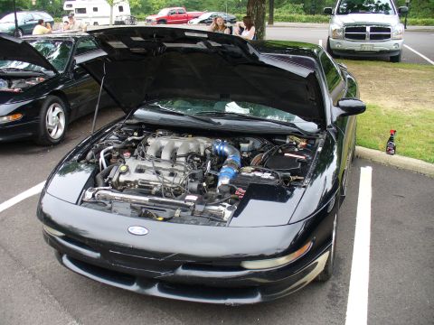 1991 Ford Probe - Engine bay of a black car is open, revealing the V6 engine and a blue intake pipe. Another car is visible to the left.