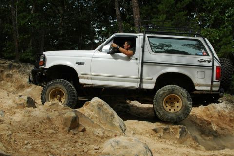 1996 Ford Bronco - A white Ford Bronco is shown off-roading on a rocky trail with trees in the background.