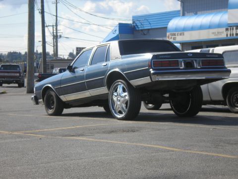 1985 Ford Bronco II - A blue Chevrolet sedan is parked in a lot, showing off its custom chrome wheels and lifted rear end.