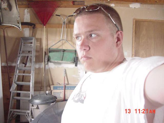 Selfie of a man in a white T-shirt inside a garage with a ladder and tools behind him