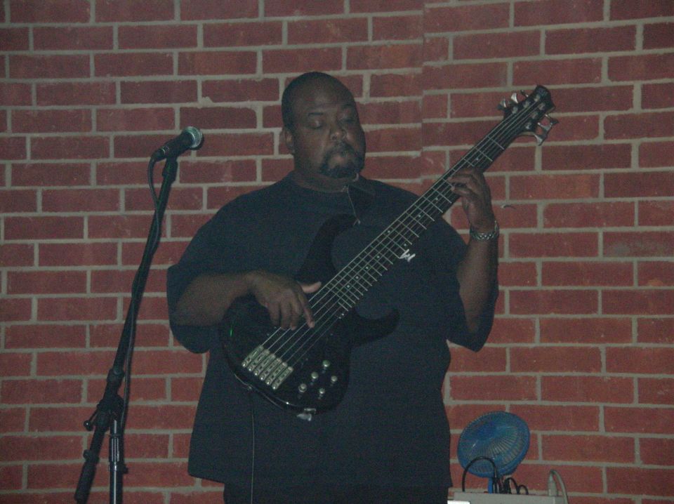 Man in a black shirt playing a five-string bass guitar in front of a brick wall, mic on a stand