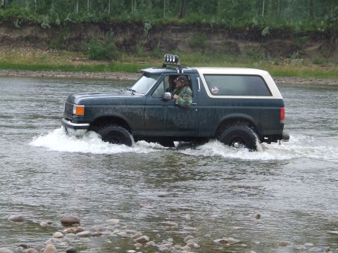 1990 Ford Bronco - A dark green Ford Bronco with a white top navigates a shallow river, kicking up water.