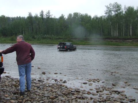 1990 Ford Bronco - A Ford Bronco navigates a shallow river crossing with a wooded bank in the background.