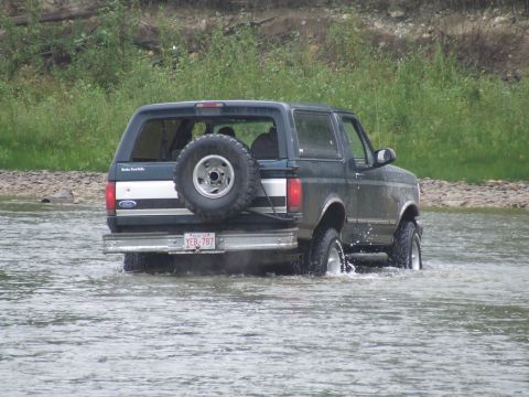 1990 Ford Bronco - A dark green 1990 Ford Bronco navigates through shallow water, kicking up spray.