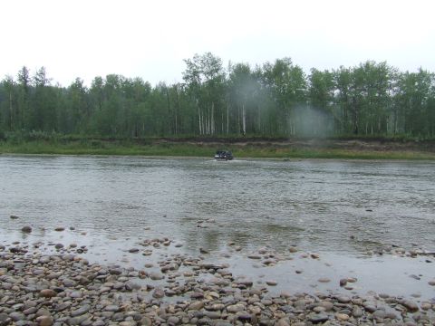 1990 Ford Bronco - A dark-colored SUV navigates across a rocky riverbed under an overcast sky.