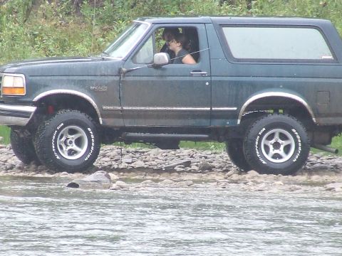 1990 Ford Bronco - A dark green 1990 Ford Bronco navigates a rocky stream bed, with a driver visible inside.