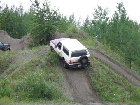 1990 Ford Bronco - A white and maroon SUV navigates a steep dirt incline, with another dark vehicle visible in the background.