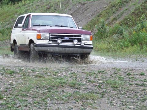 1990 Ford Bronco - A two-tone Ford Bronco splashes through water on a gravelly path.