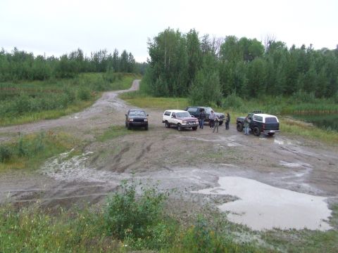 1990 Ford Bronco - A group of off-road vehicles are gathered on a dirt path near a body of water, surrounded by dense forest.