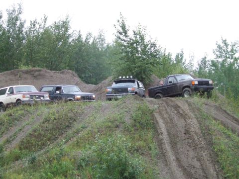 1990 Ford Bronco - Four Ford SUVs and trucks are parked on a dirt hill with trees in the background.