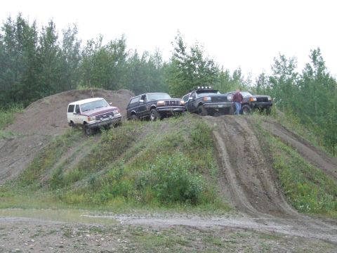 1990 Ford Bronco - Several SUVs, including a 1990 Ford Bronco, navigate a muddy dirt hill in an off-road setting.