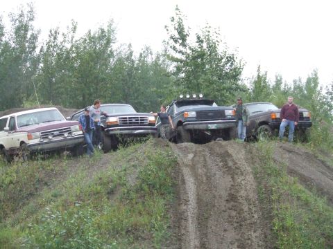 1990 Ford Bronco - Several off-road vehicles and their occupants are gathered on a dirt embankment under an overcast sky.