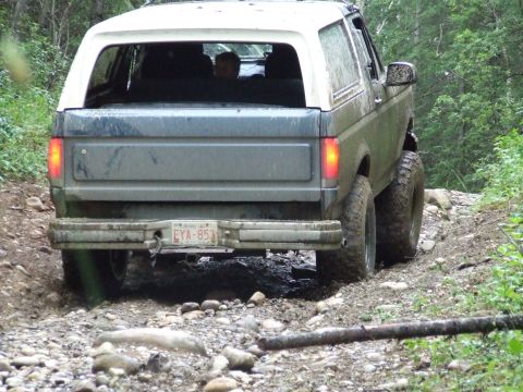 1990 Ford Bronco - A 1990 Ford Bronco navigates a rough, wooded path, its rear taillights illuminated.