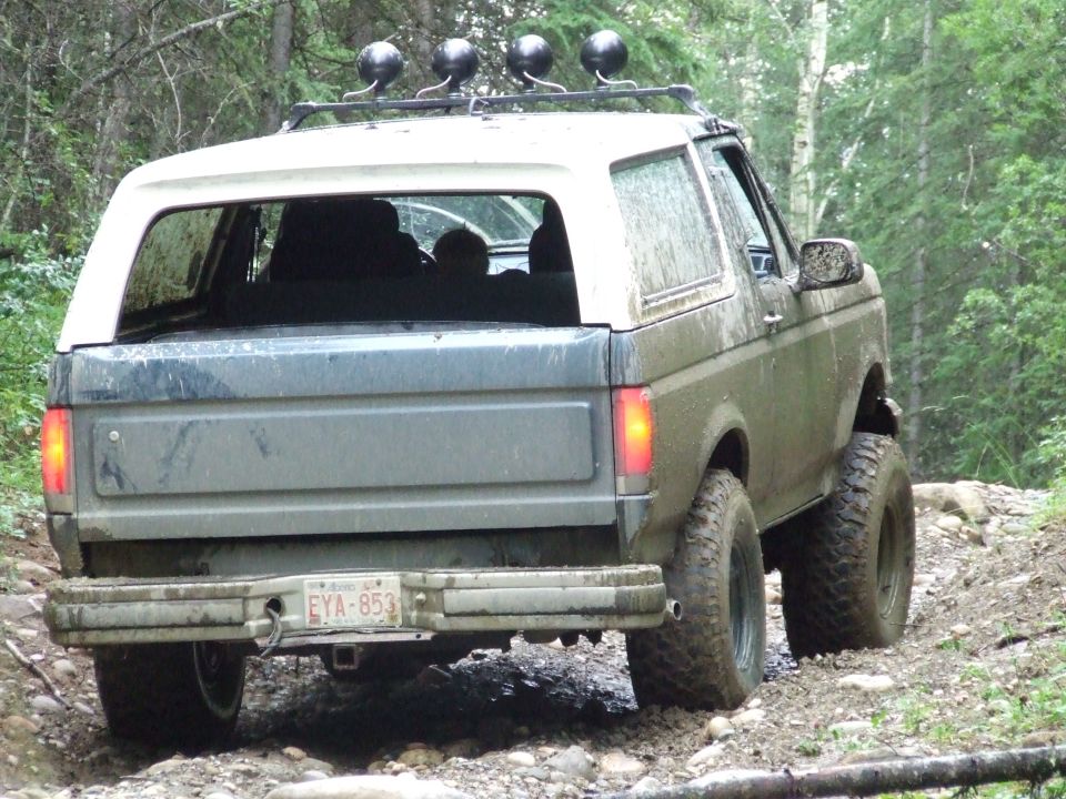 Mud-splattered 1987 Ford Bronco, rear view, off-road tires, roof rack with lights, light-colored cap