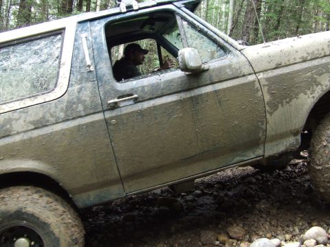 1990 Ford Bronco - A heavily mud-splattered blue Ford Bronco navigates a rocky, off-road path with its driver behind the wheel.