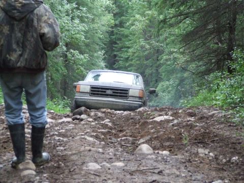 1990 Ford Bronco - A person in camouflage stands on a rough forest path as a Ford Bronco drives through mud and rocks.