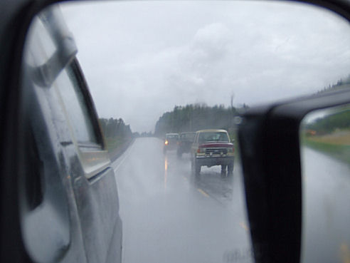 Red and tan SUV driving in rain, seen in side mirror