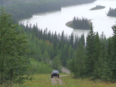 1988 Ford Bronco - A lone SUV navigates a muddy track amidst a dense forest, with a wide river winding through the landscape.