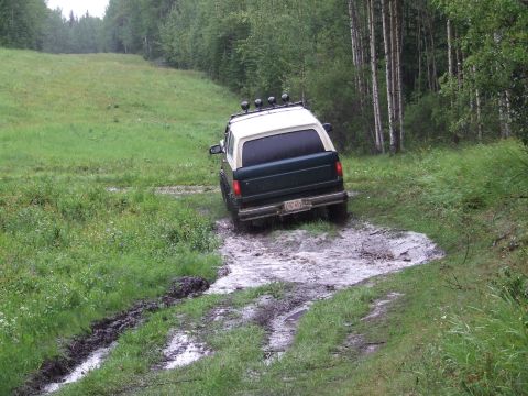 1988 Ford Bronco - Rear view of a green and white full-size Ford Bronco pushing through a wide mud hole on a grassy trail, throwing muddy water off its tires.…