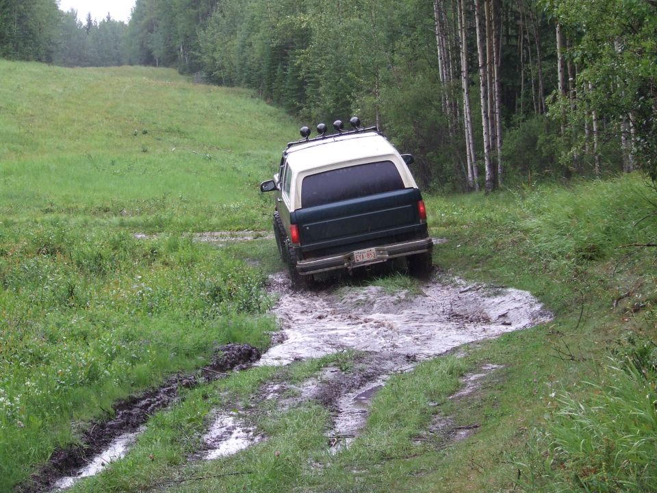 Green and white Ford Bronco churning through a mud hole, viewed from the rear