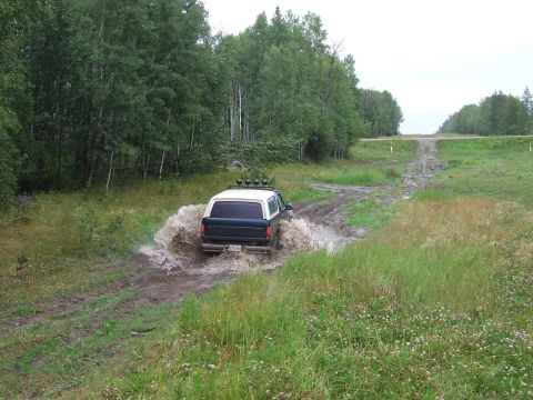 1988 Ford Bronco - An SUV navigates a muddy trail, kicking up a large splash as it drives through a water-filled rut.