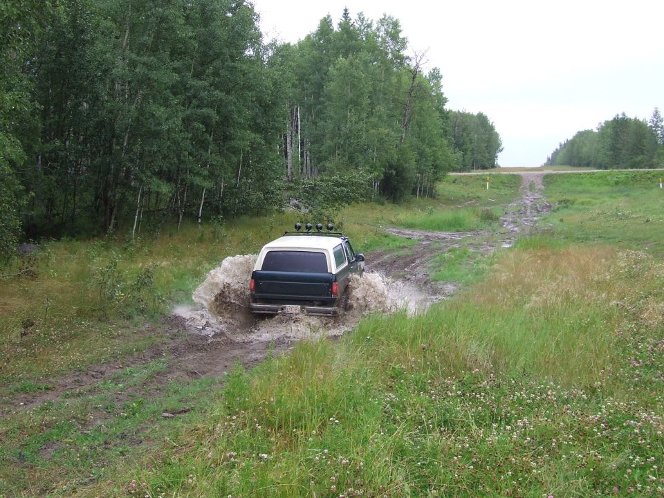 SUV splashing through a muddy puddle on a dirt road, trees and sky in background