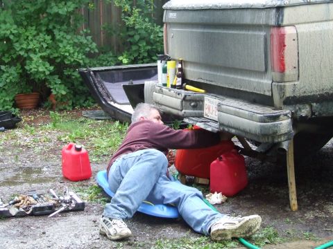 1990 Ford Bronco - A man works on the underside of a pickup truck in a yard, with tools and gas cans scattered around.