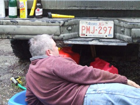 1990 Ford Bronco - Man working under vehicle bumper with gas cans visible. Alberta license plate PMJ-297.