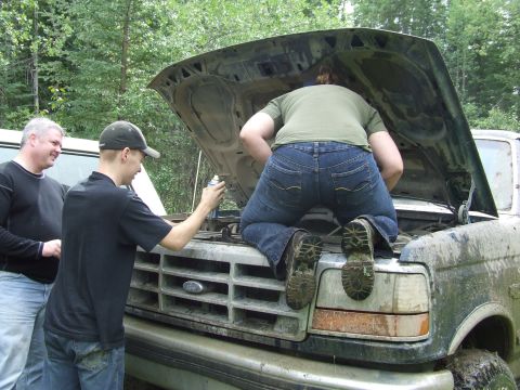 1990 Ford Bronco - Three people attend to a dirty Ford Bronco with its hood up, suggesting mechanical work in an outdoor setting.