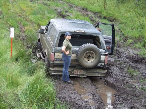 1990 Ford Bronco - A woman poses next to a muddy Ford Bronco that is stuck off-road.