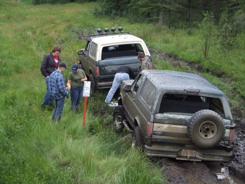 1990 Ford Bronco - Two Ford Broncos are stuck in the mud on a trail with several people observing.
