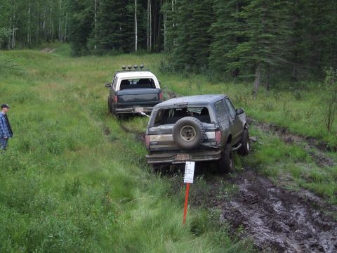 1990 Ford Bronco - Two Ford Broncos navigate a muddy off-road trail surrounded by trees.