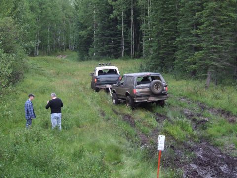 1990 Ford Bronco - Two vehicles navigate a muddy trail through a forest, with two people observing the scene.