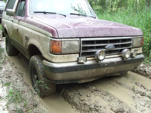 1990 Ford Bronco - A maroon and tan 1990 Ford Bronco navigates a muddy track, its front tires deep in the mire.