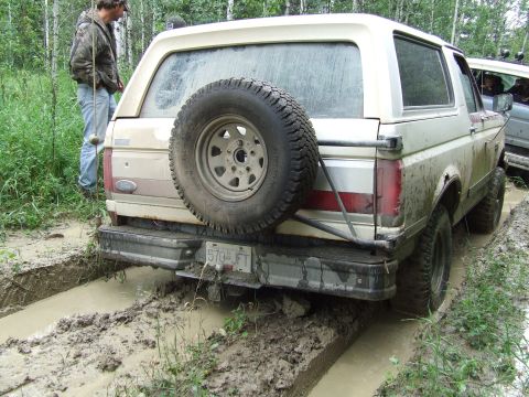 1990 Ford Bronco - A tan Ford Bronco navigates a muddy track in a wooded area, with another vehicle visible to the right.
