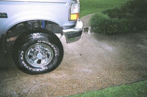1995 Ford Bronco - The front quarter panel and tire of a silver Ford Bronco are visible on a wet driveway.