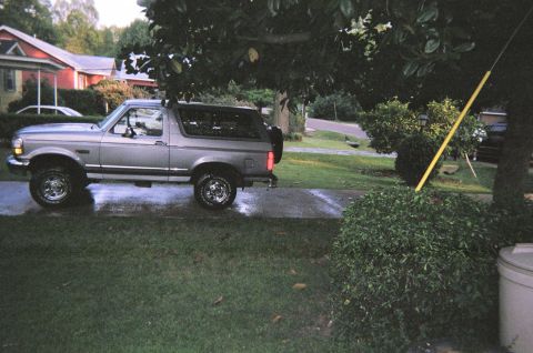 1995 Ford Bronco - Silver 1995 Ford Bronco parked on a wet driveway next to a lawn and trees.