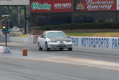 1992 Ford Taurus - A white 1992 Ford Taurus with a black hood drives down a drag strip, passing a starting light tree.