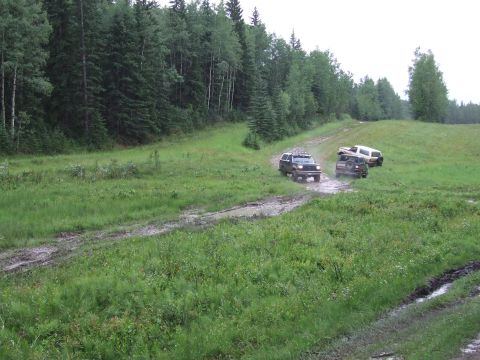 1988 Ford Bronco - Multiple SUVs drive off-road through a muddy trail with trees in the background.